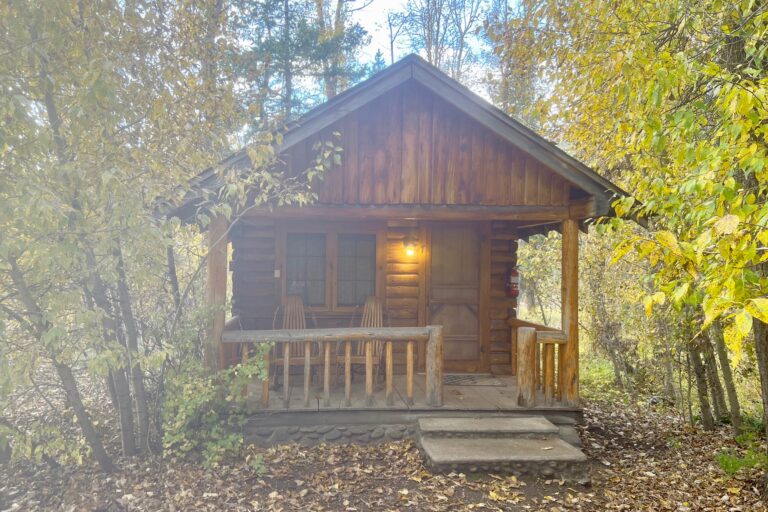 The front view of a cabin shows trees nearby and a spacious porch.