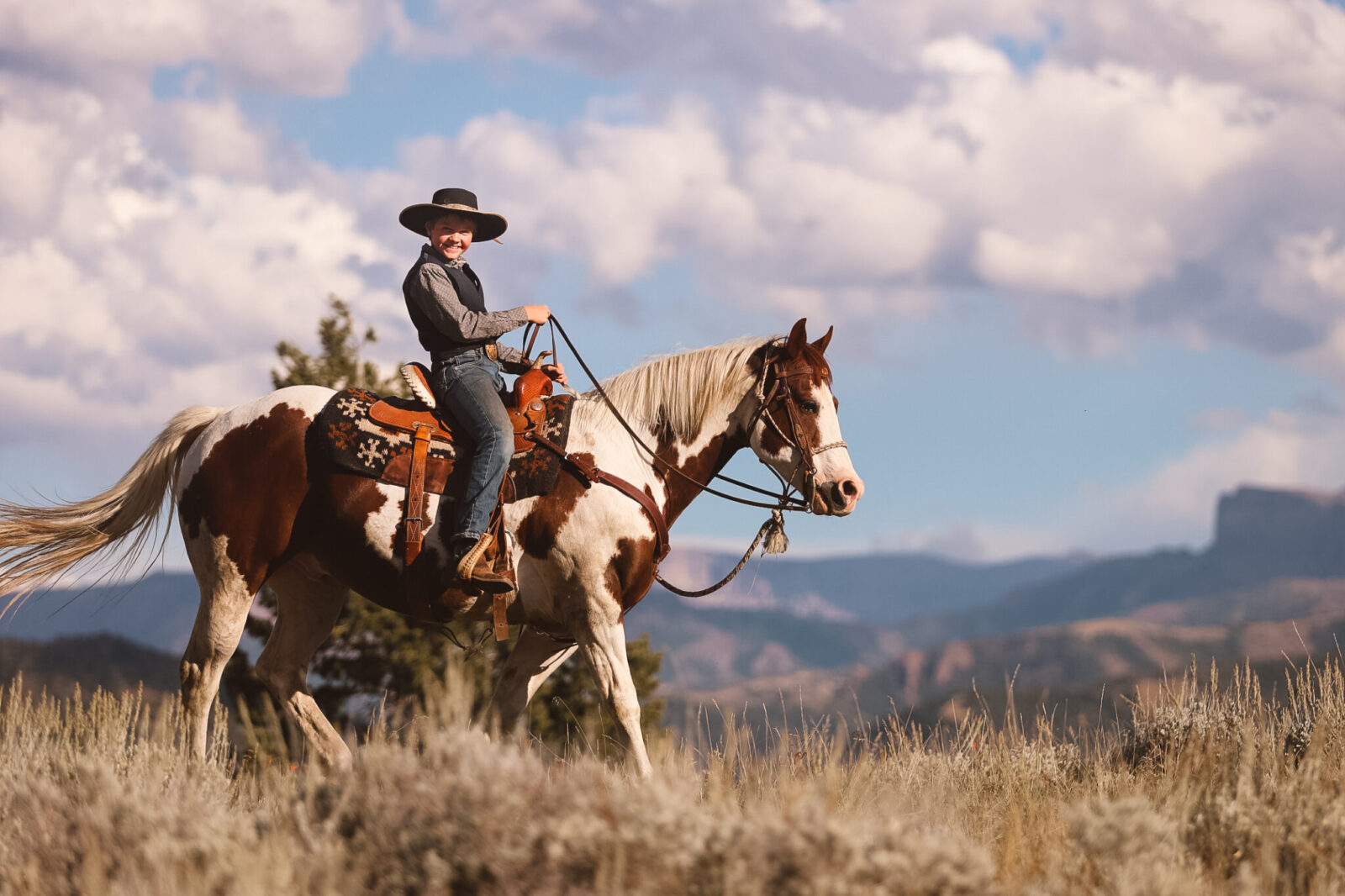 A smiling boy in a black cowboy hat rides a stocky Paint gelding in front of a grand mountain view.