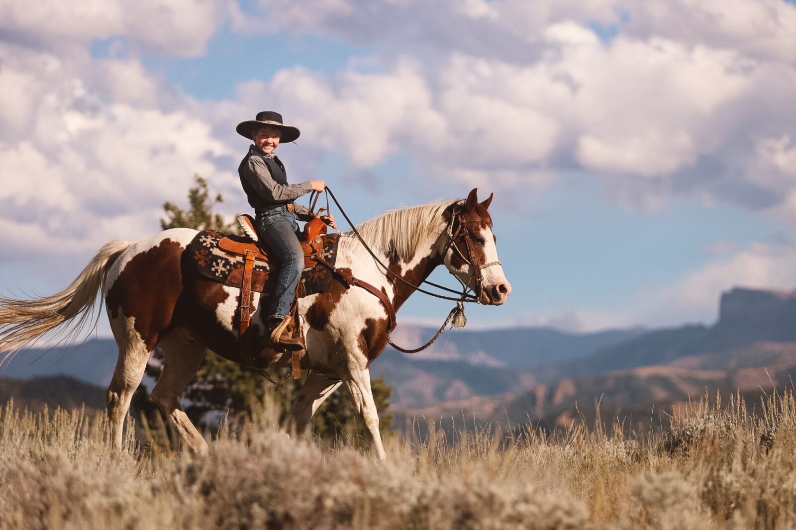 Cade A smiling boy in a black cowboy hat rides a stocky Paint gelding in front of a grand mountain view.