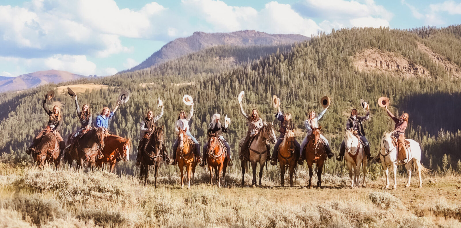 A group of cowgirls on horses hold their hats in the air in celebration with a lovely mountain scene as the backdrop.