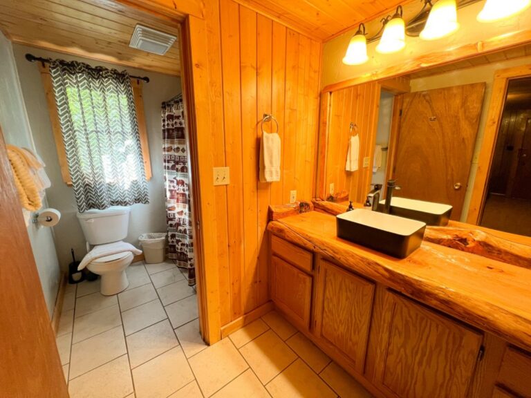 Rustic cabin bathroom with knotty pine walls, live-edge wood vanity, black vessel sink, and toilet beside a curtained window.
