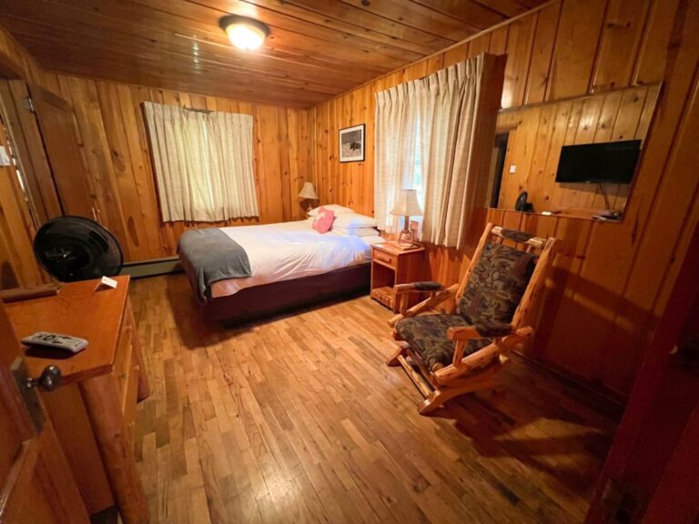 Knotty pine-paneled cabin bedroom with a queen bed, log rocking chair, wall-mounted TV, and hardwood floor.