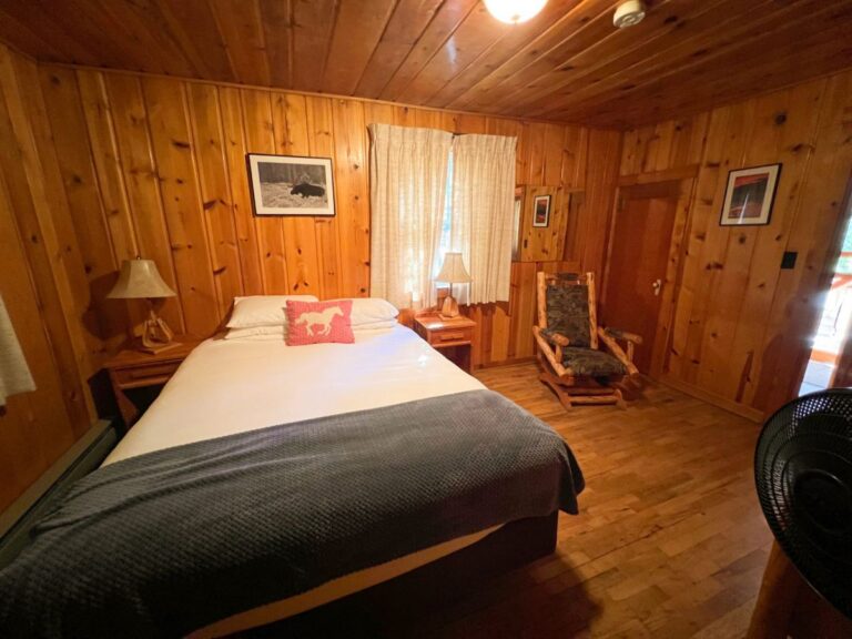 Rustic knotty-pine cabin bedroom with a queen bed, red horse-print pillow, log rocking chair, and curtained window.