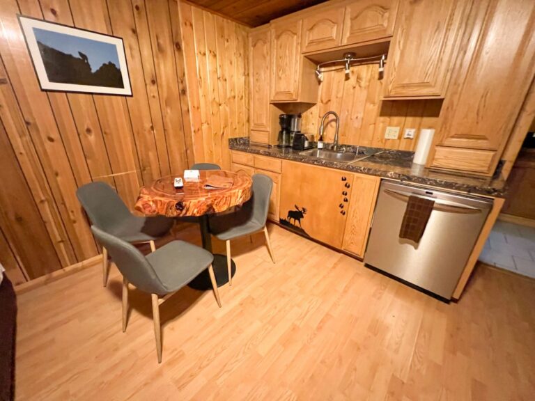 Small kitchenette in a knotty pine cabin with a live-edge wood table, two gray chairs, stainless dishwasher, and a moose cutout on the cabinet.