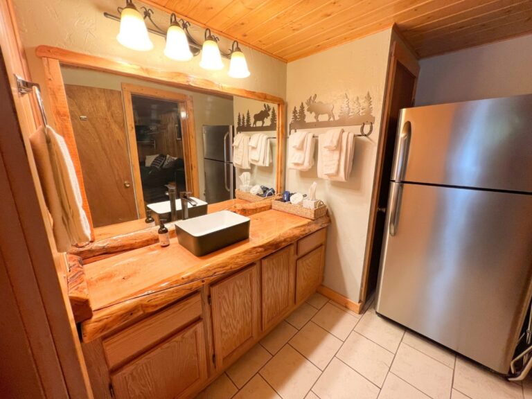 Rustic cabin bathroom with a live-edge wood vanity, vessel sink, moose and pine wall decor, white towels, and a stainless steel refrigerator.