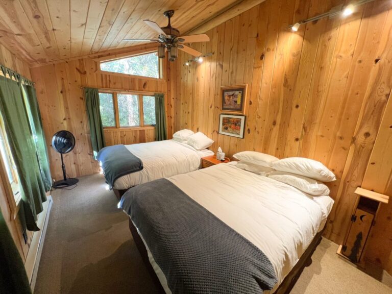 Rustic wood-paneled cabin bedroom with two beds in white linens and gray throws, ceiling fan, and forest-view windows.