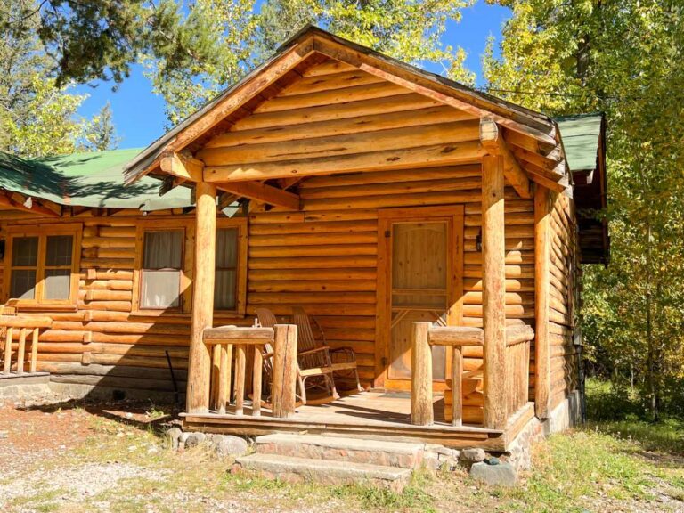 Rustic log cabin with a green metal roof, covered front porch, wooden rocking chairs, and stone steps, surrounded by sunlit aspen and pine trees.