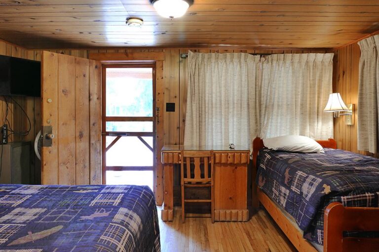 Rustic knotty pine cabin room with two twin beds in blue plaid quilts, a log desk and chair, and a screen door.